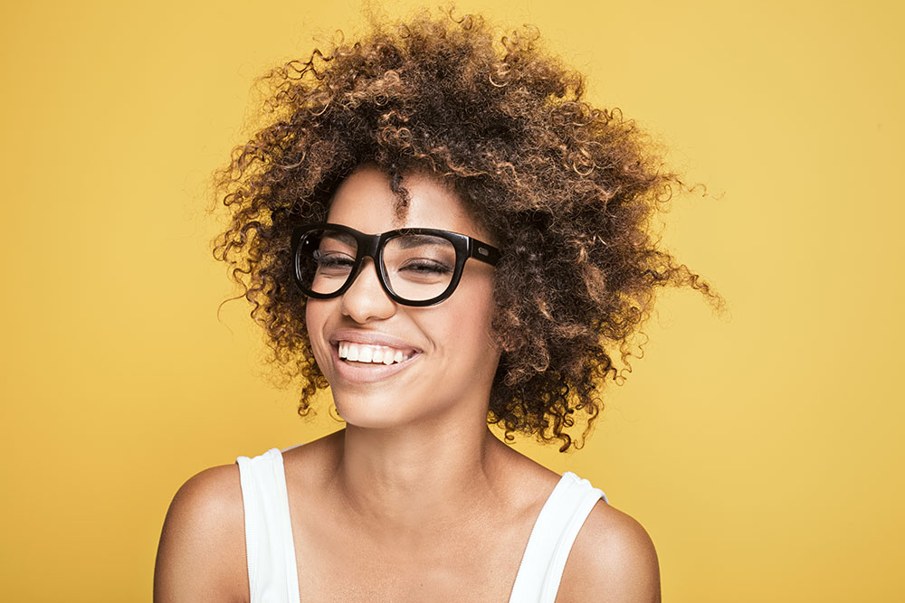 A woman with a wide smile, curly hair, and wearing dark glasses, sitting with an orange background